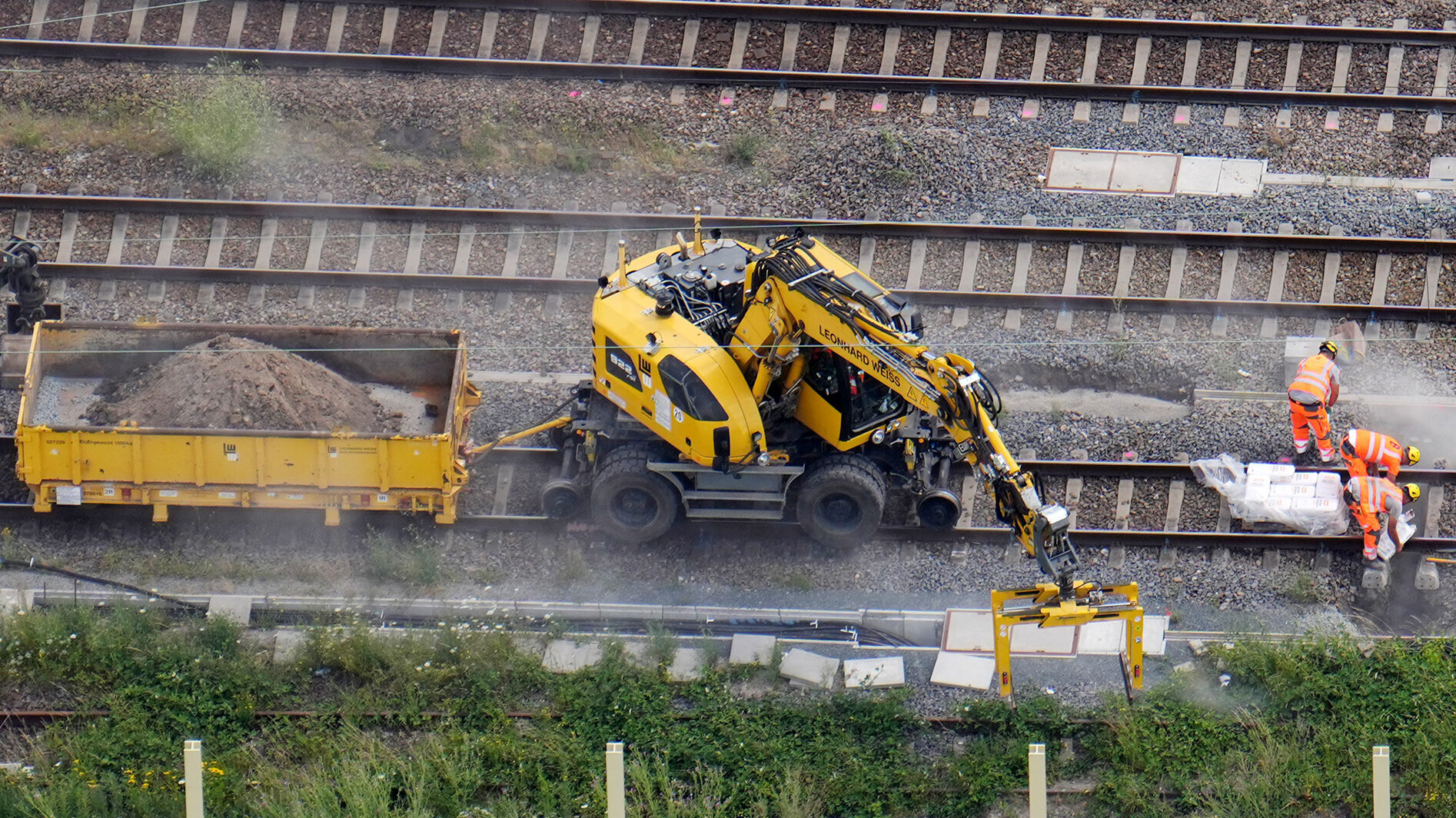 Auf der Baustelle sind mehrere Arbeiter beschäftigt, während ein schweres Gerät Erde in einen Lkw lädt.