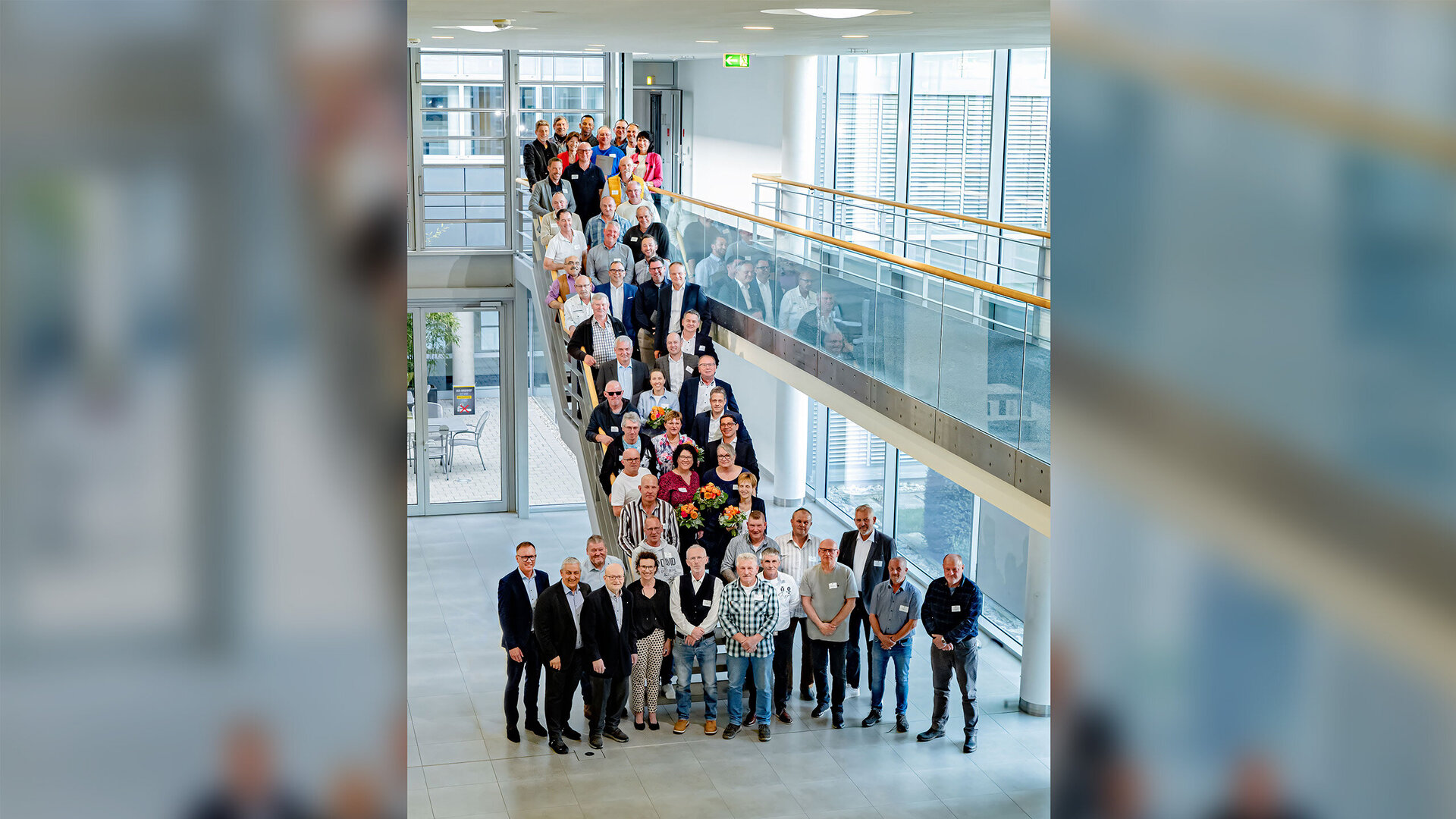 Große Gruppe von Mitarbeitenden steht auf einer Treppe in einem modernen, hellen Firmengebäude und blickt in die Kamera. Einige Personen halten Blumensträuße, was auf eine Ehrung oder ein Jubiläum hinweist.