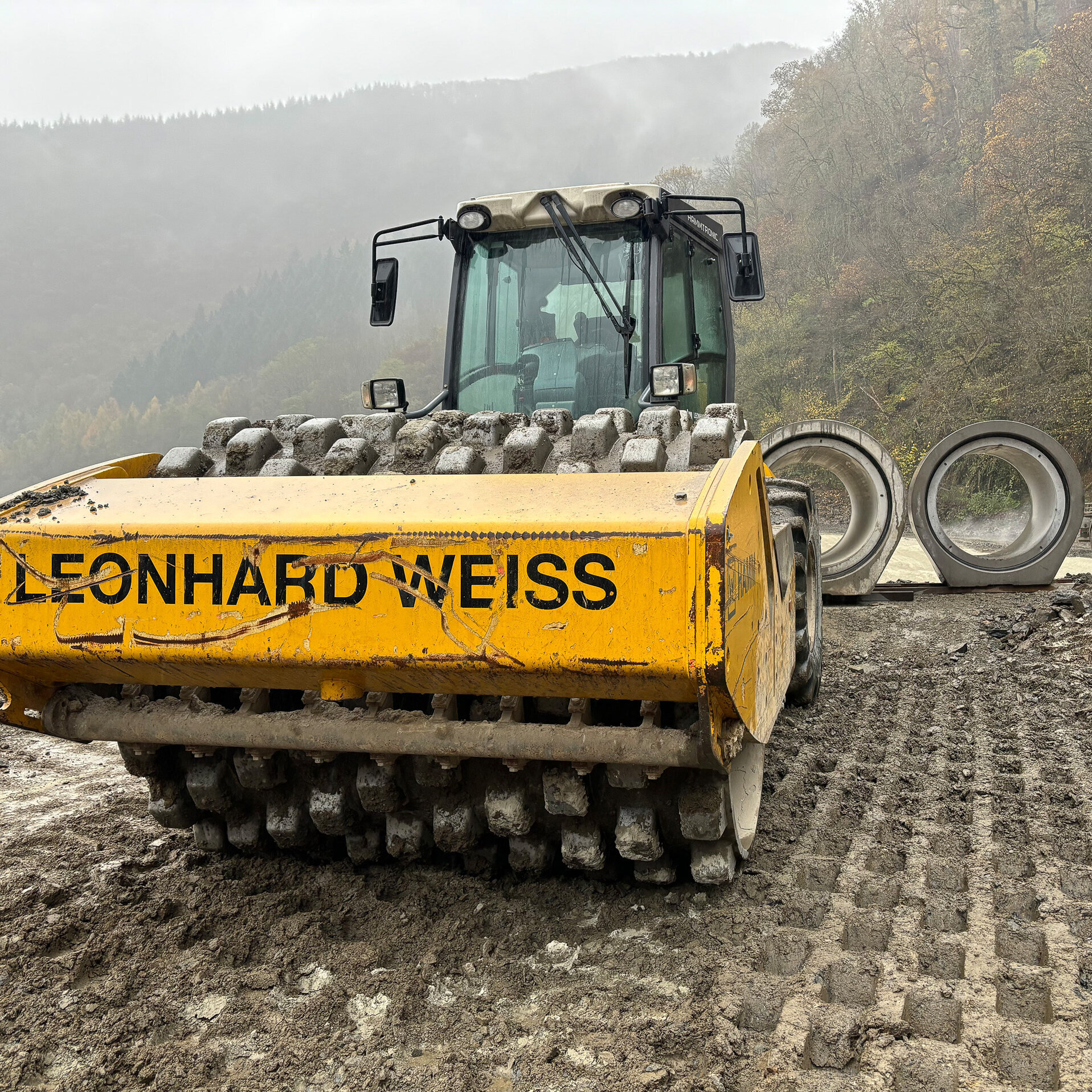 Ein gelber Walzenfahrer steht im Vordergrund, während große Betonrohre im Hintergrund auf einer Baustelle liegen.