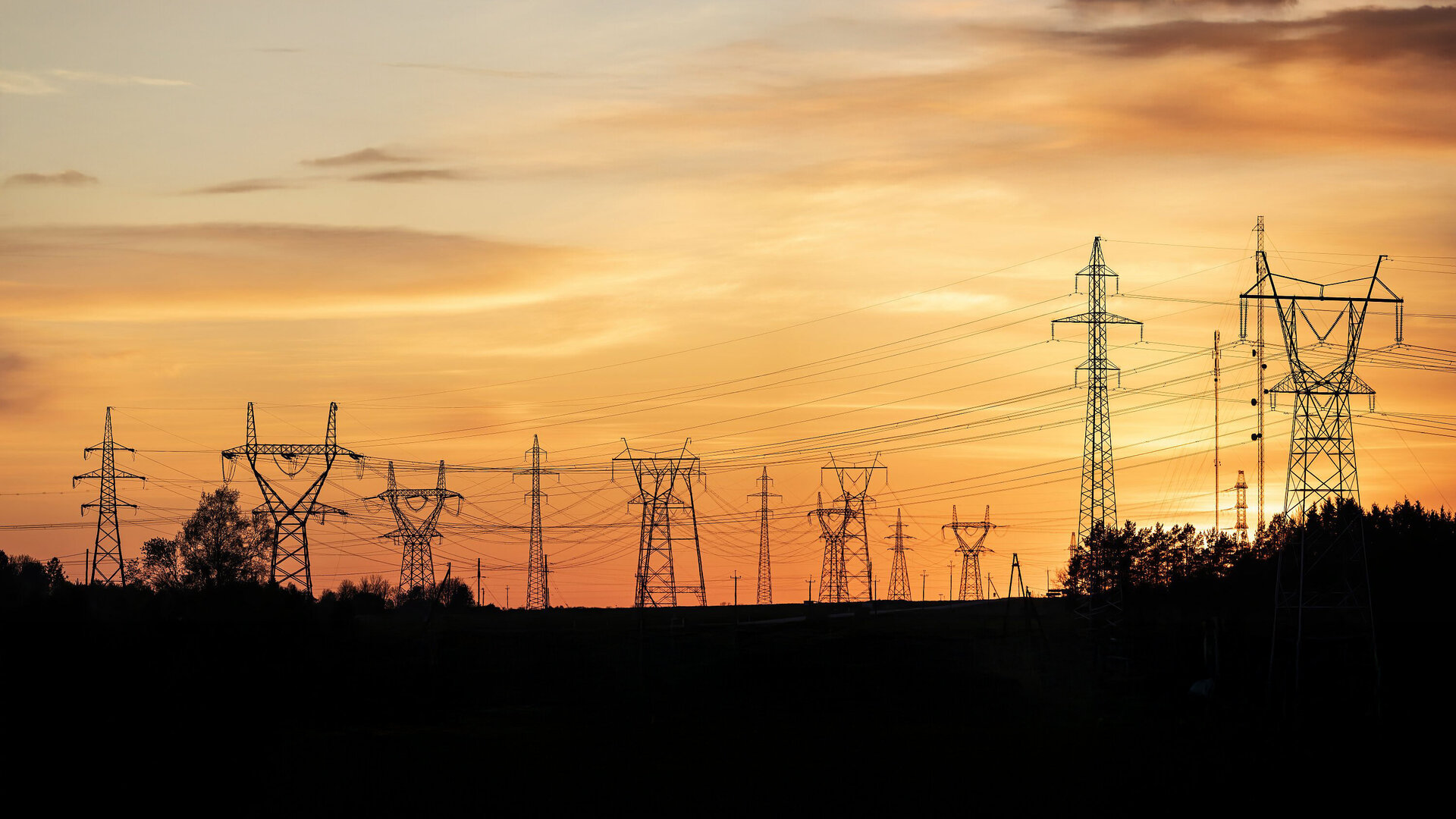 High-voltage power lines stretch across the landscape as the sun sets behind the horizon, lighting up the sky.