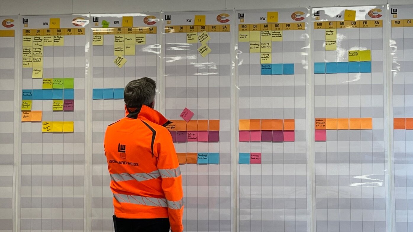 In einem modernen Büro analysiert ein Mann in Sicherheitskleidung die Fortschritte auf einem großen, strukturierten Planungsboard.