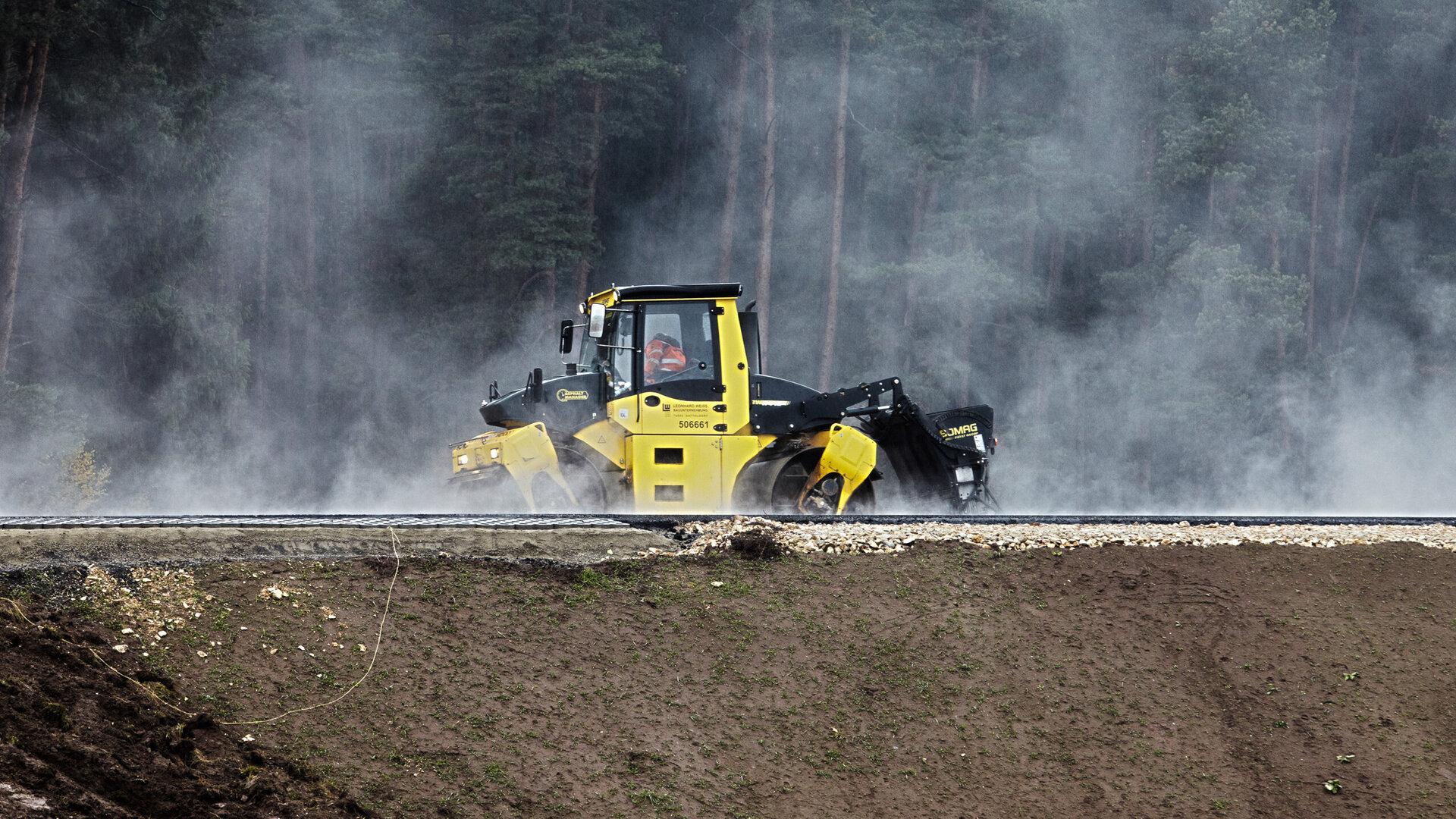 The machine, surrounded by mist, works the soil while trees are visible in the background, framing the scene.