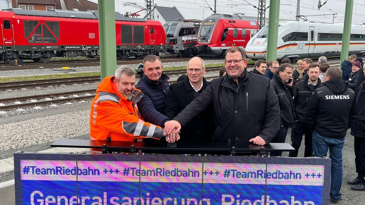 Eine Gruppe von Männern posiert vor einem großen Bildschirm, der die Wiederinbetriebnahme der Riedbahn verkündet, umgeben von Zügen.
