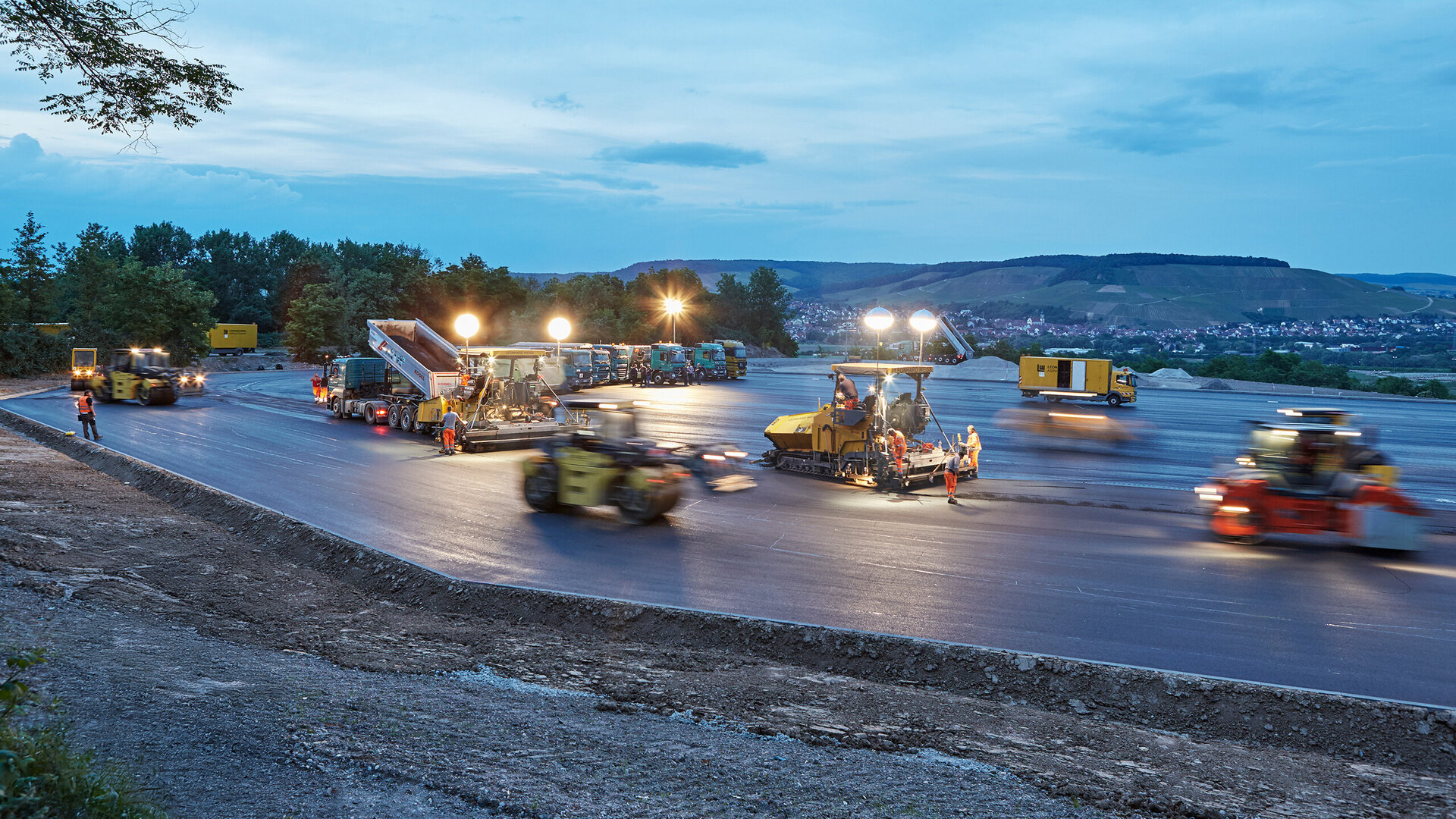 Bei Dämmerung arbeiten Bauarbeiter an einer neuen Straße, umgeben von schweren Maschinen und hellen Scheinwerfern.