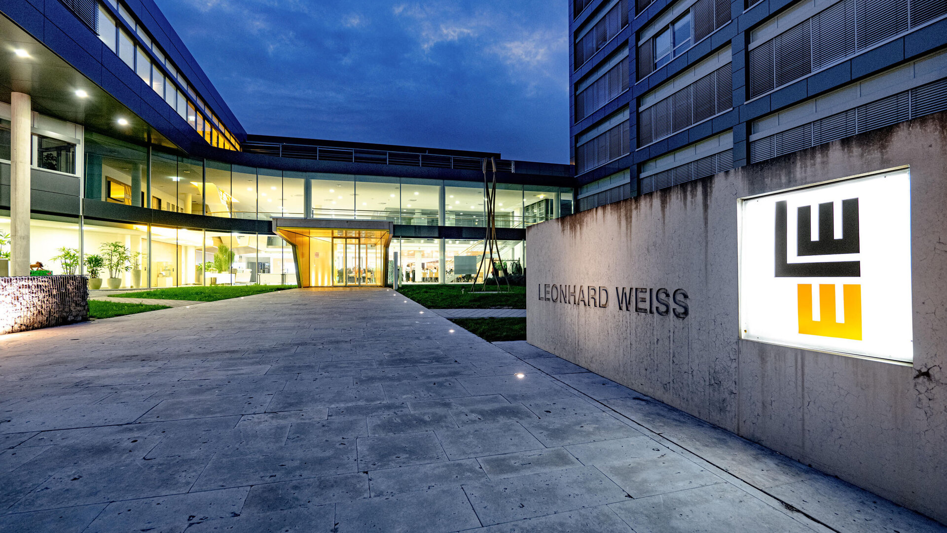 Abendaufnahme des modernen Firmengebäudes von Leonhard Weiss mit beleuchtetem Eingang und großem Firmenlogo an einer Steinwand. Durch die Glasfront ist der helle, einladende Innenbereich sichtbar.