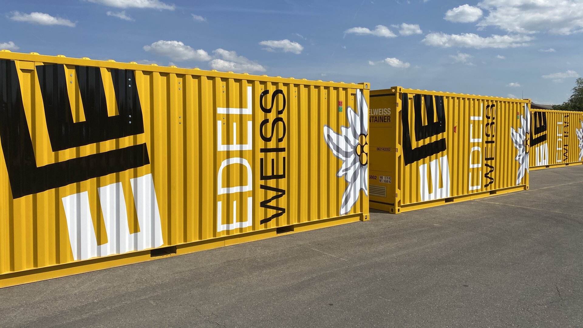 Die gelben Container sind mit dem Schriftzug 'EDELWEISS' und einer stilisierten Edelweißblume dekoriert, unter einem blauen Himmel.