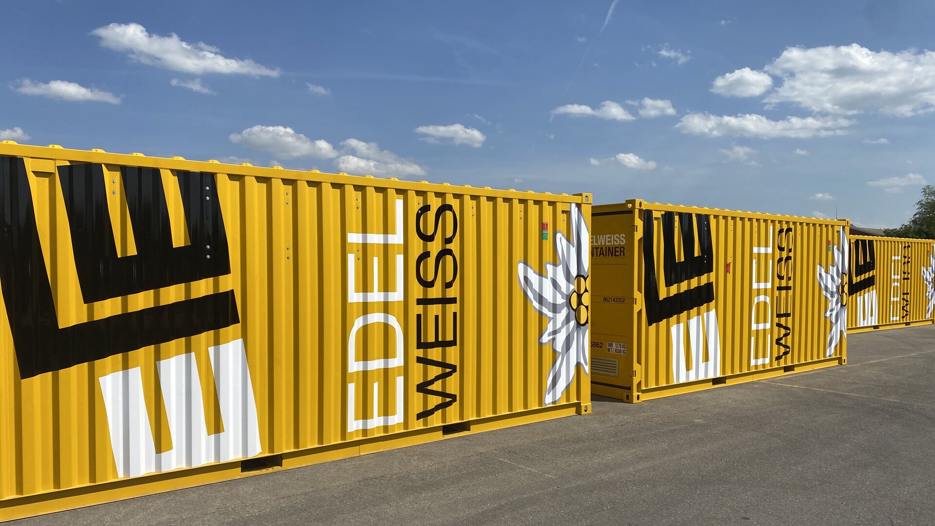 Die gelben Container sind mit dem Schriftzug 'EDELWEISS' und einer stilisierten Edelweißblume dekoriert, unter einem blauen Himmel.
