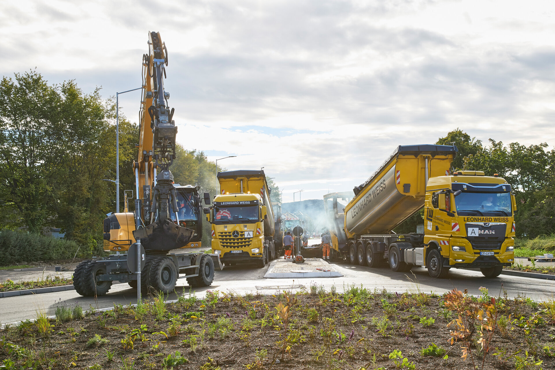 Auf einer Baustelle stehen zwei gelbe Lkw und ein Bagger, während Arbeiter an der Straße arbeiten und Rauch aufsteigt.