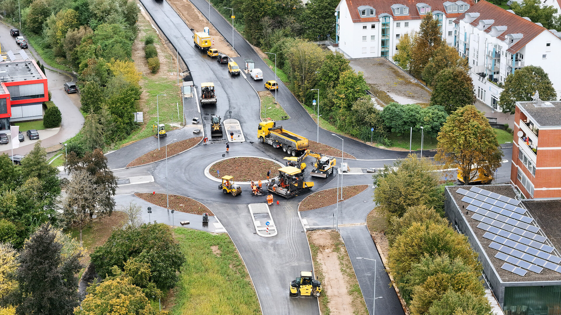 Eine Baustelle mit mehreren gelben Baumaschinen, die an einem neuen Kreisverkehr arbeiten, umgeben von Bäumen und Wohngebäuden.