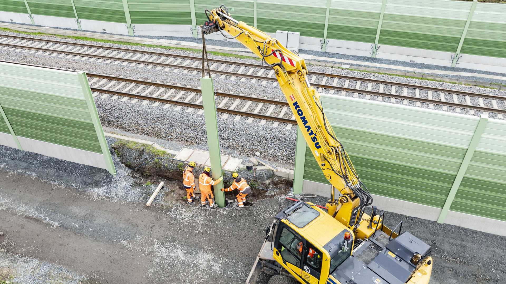 Fünf Bauarbeiter arbeiten an einer Baustelle neben einer Bahngleisanlage, während ein Bagger einen Pfosten anhebt.