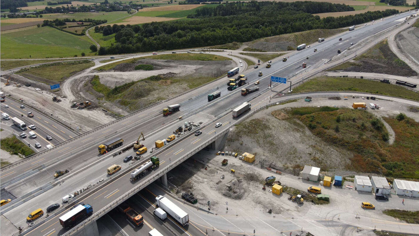 Eine Luftaufnahme zeigt eine vielbefahrene Baustelle mit mehreren Baustellenfahrzeugen und Lkw auf einer Autobahn.