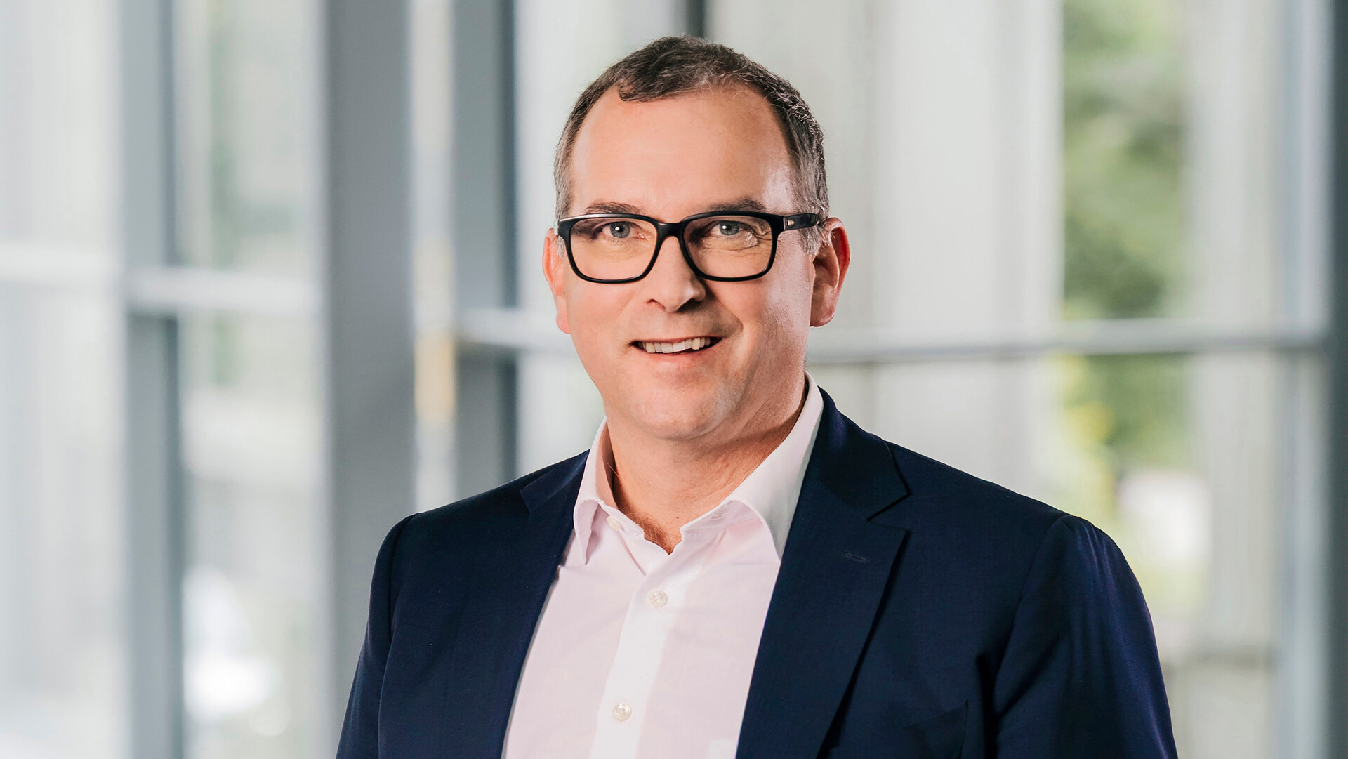 Foto von Alexander Weiss, Ressortleiter Personal und Kommunikation, stehend in Anzug und Hemd in einem modernen Bürogebäude.