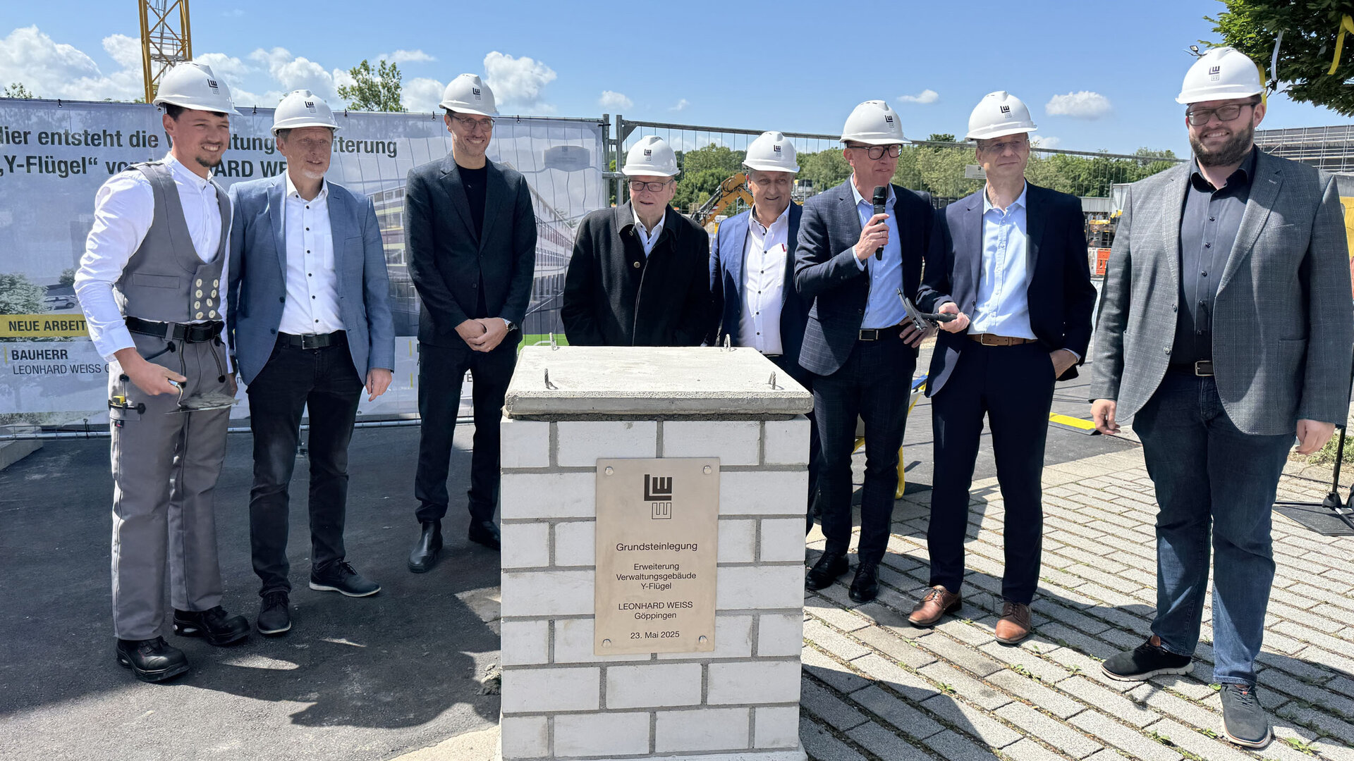 Die Anwesenden, gekleidet in Anzügen und Baukleidung, feiern den Grundstein für ein neues Bauprojekt an einem sonnigen Tag.