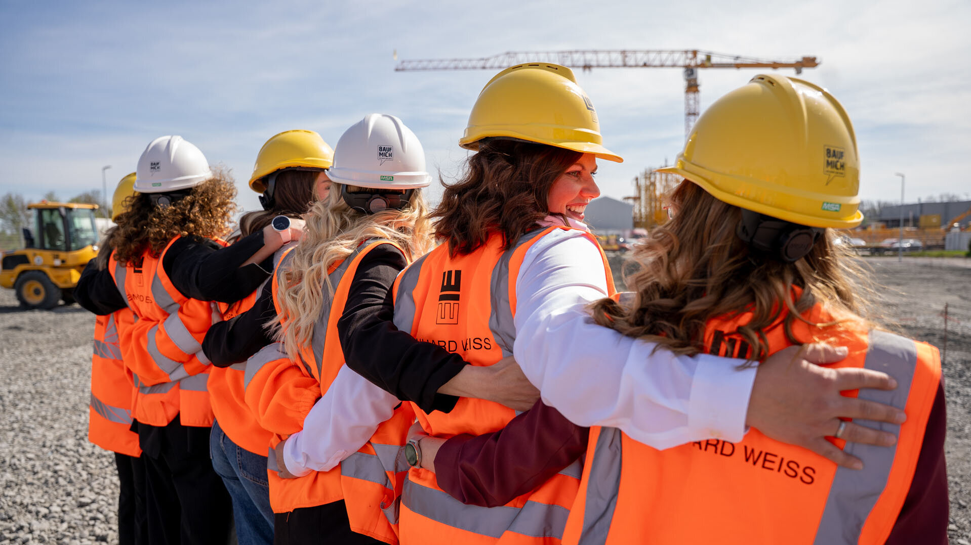 Auf einer Baustelle stehen Frauen in Sicherheitskleidung, die sich umarmen und den Zusammenhalt in ihrem Team demonstrieren.