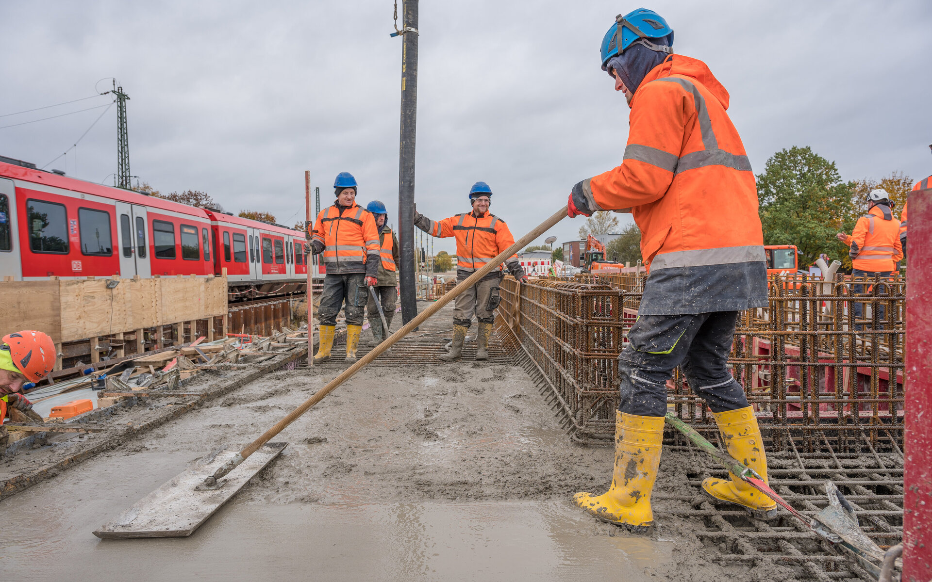 Fünf Bauarbeiter in orangefarbenen Sicherheitsjacken arbeiten an Betonage-Decke Personenunterführung mit Aufkantung für das Oberlicht, während ein Zug im Hintergrund vorbeifährt.