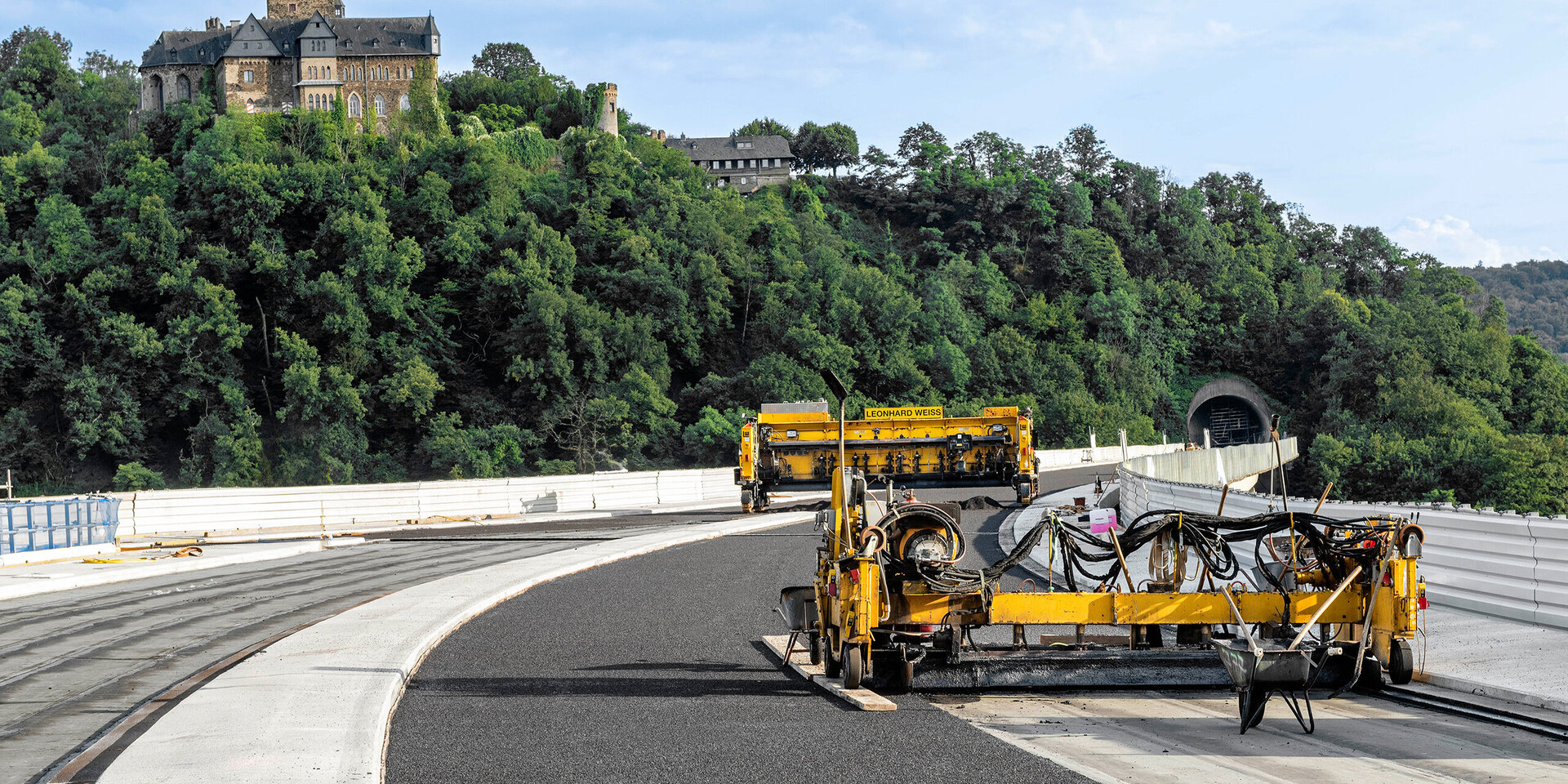 Eine Baustelle zeigt gelbe Maschinen, die Asphalt auf einer kurvenreichen Straße verlegen, mit einer historischen Burg im Hintergrund.