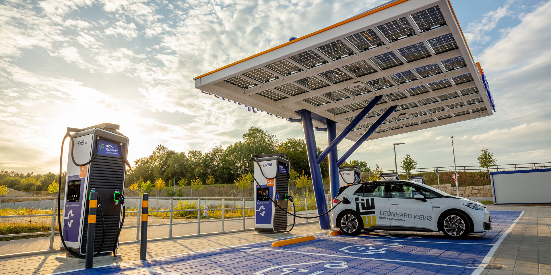 Ein Elektrofahrzeug parkt an einer innovativen Ladestation, die von Sonnenlicht gespeist wird, umgeben von Bäumen und einem blauen Himmel.