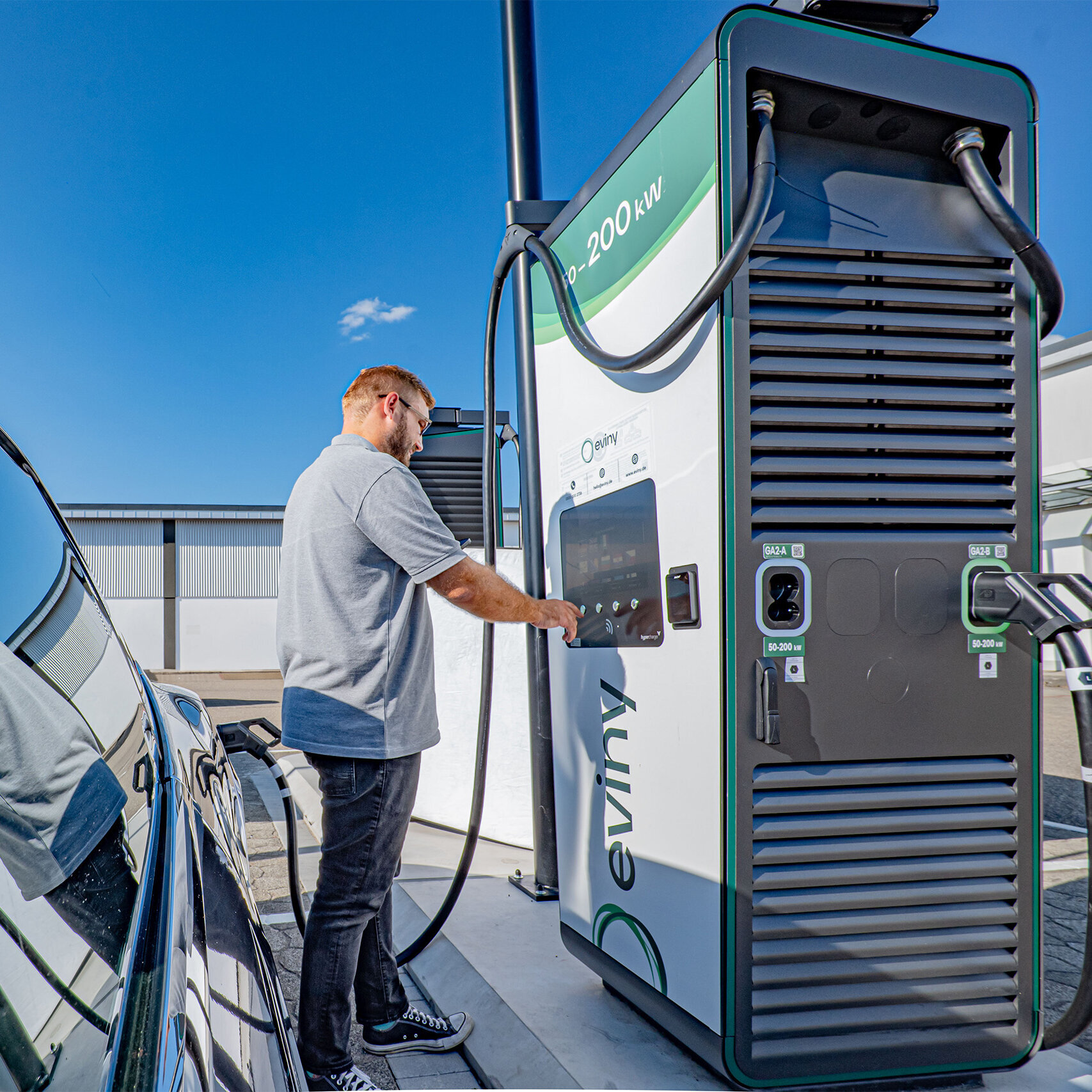 Der Fahrer eines Elektroautos interagiert mit einer modernen Ladestation, um sein Fahrzeug aufzuladen, bei klarem Himmel.