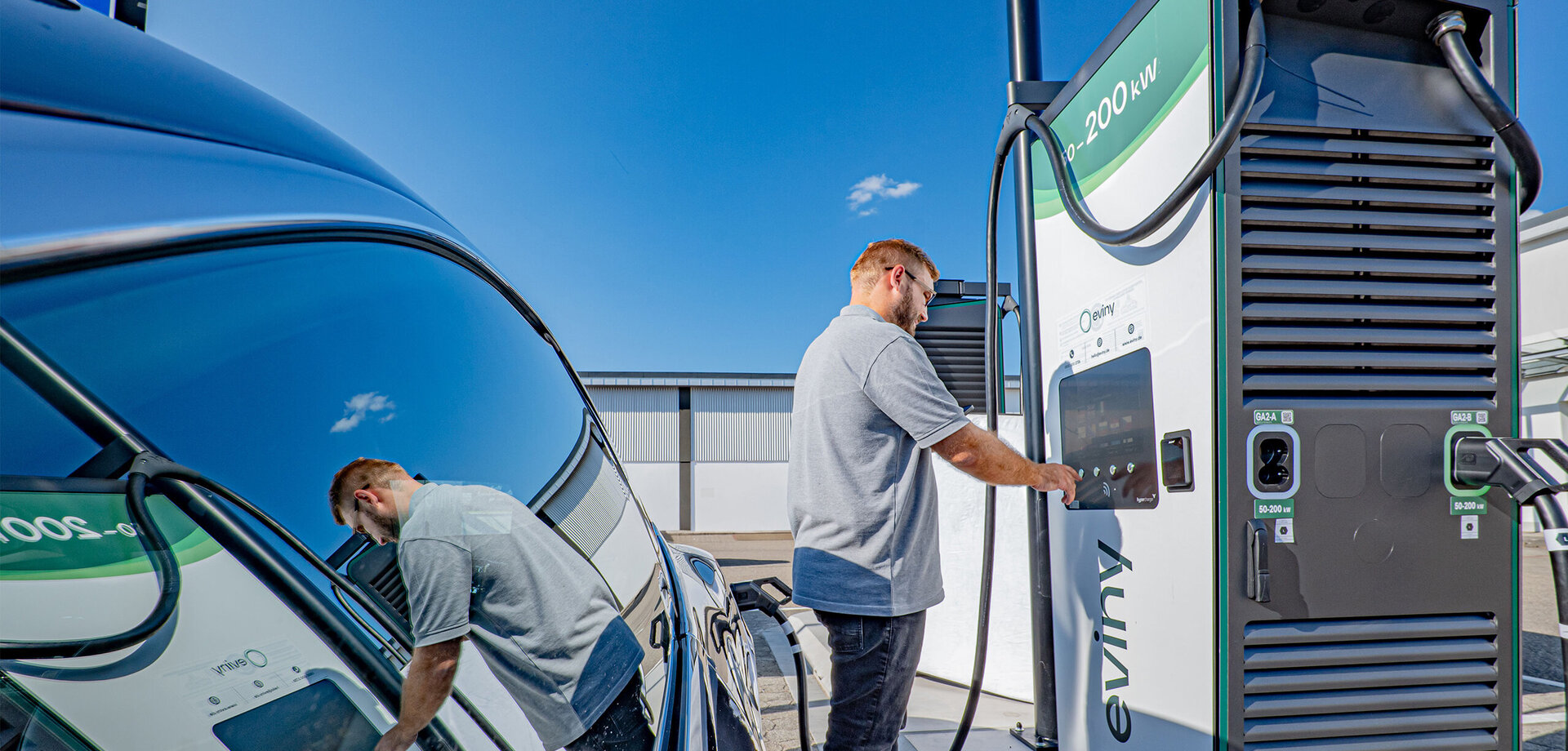 Der Fahrer eines Elektroautos interagiert mit einer modernen Ladestation, um sein Fahrzeug aufzuladen, bei klarem Himmel.