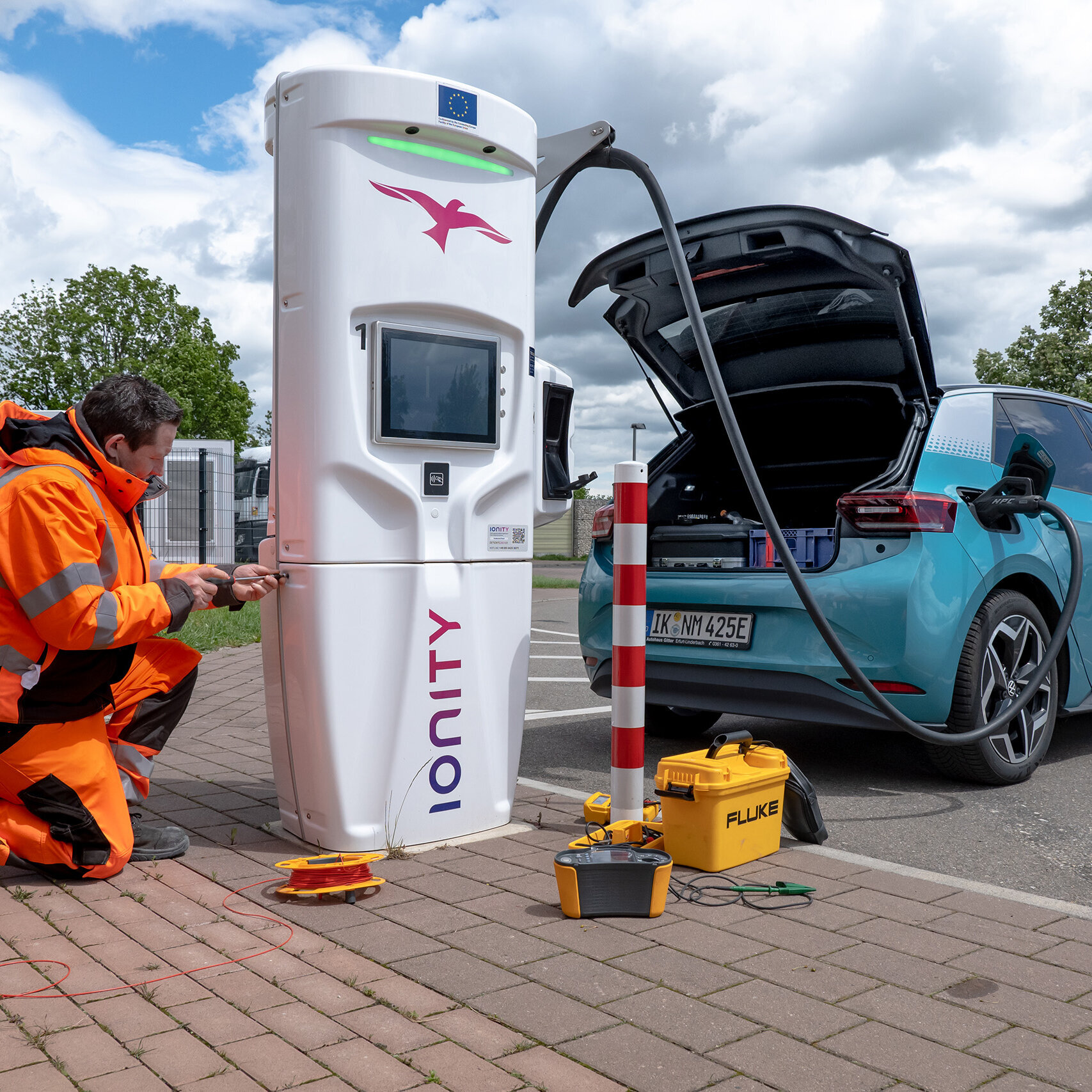 Ein Mann kniet vor einer Ionity-Ladestation und arbeitet daran, während ein Elektroauto im Hintergrund aufgeladen wird.
