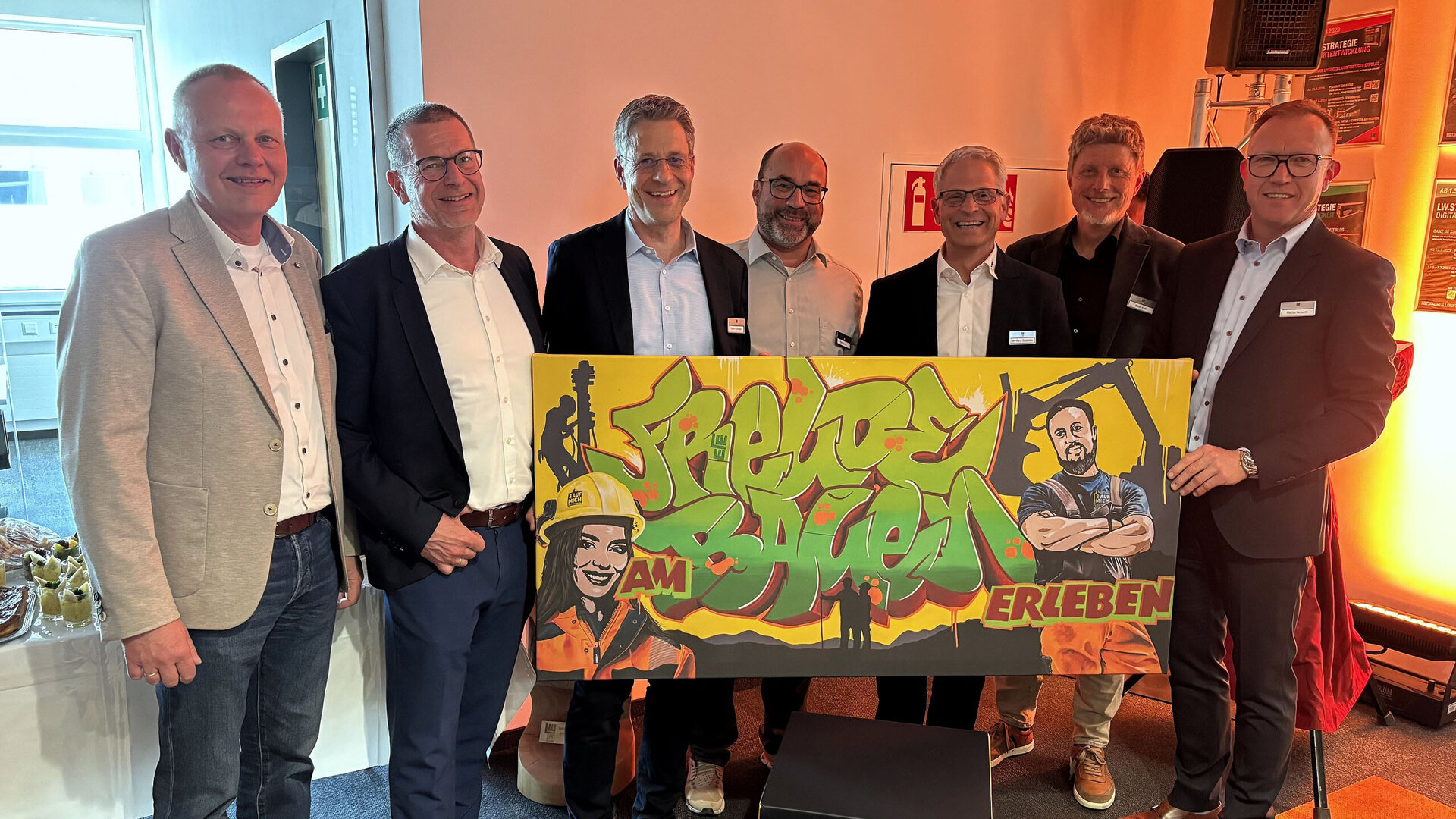 Sechs Männer in Anzügen stehen zusammen und halten ein großes, buntes Plakat mit Graffiti-Design in der Hand.