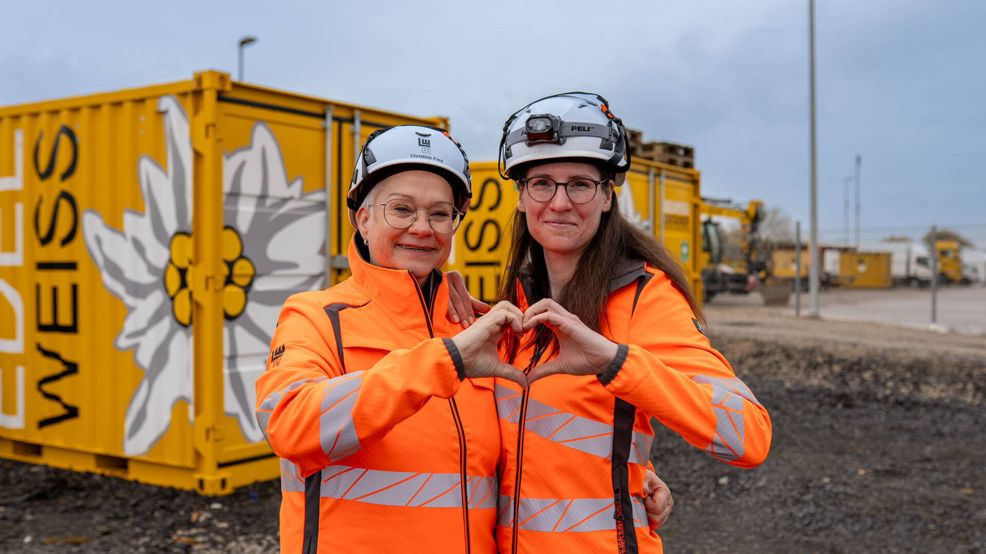 Zwei Frauen in leuchtend orangefarbener Arbeitskleidung formen ein Herz mit ihren Händen vor gelben Containern.