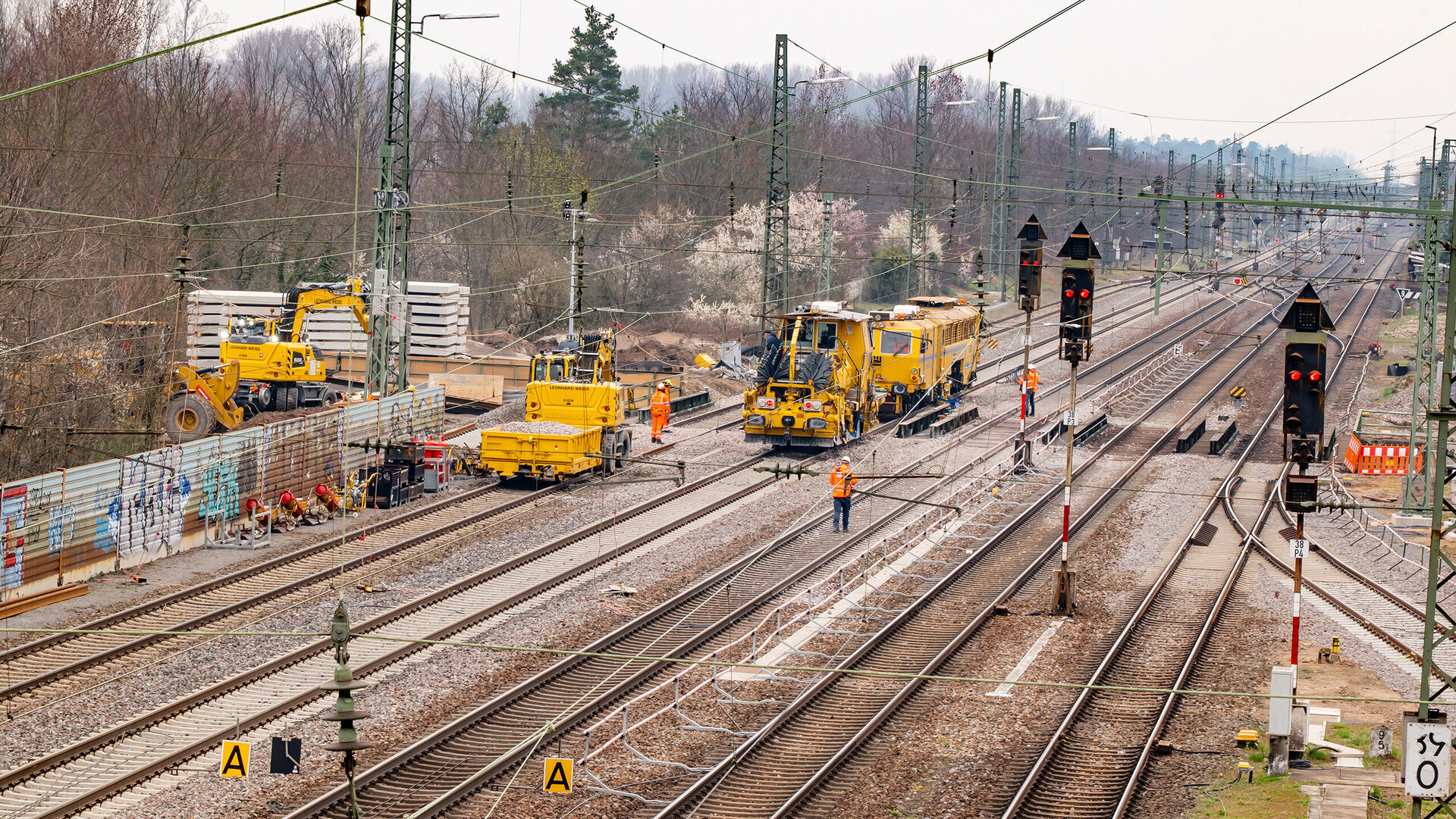 Auf einer Baustelle arbeiten mehrere gelbe Maschinen an den Gleisen, während Arbeiter in orangefarbener Kleidung aktiv sind.