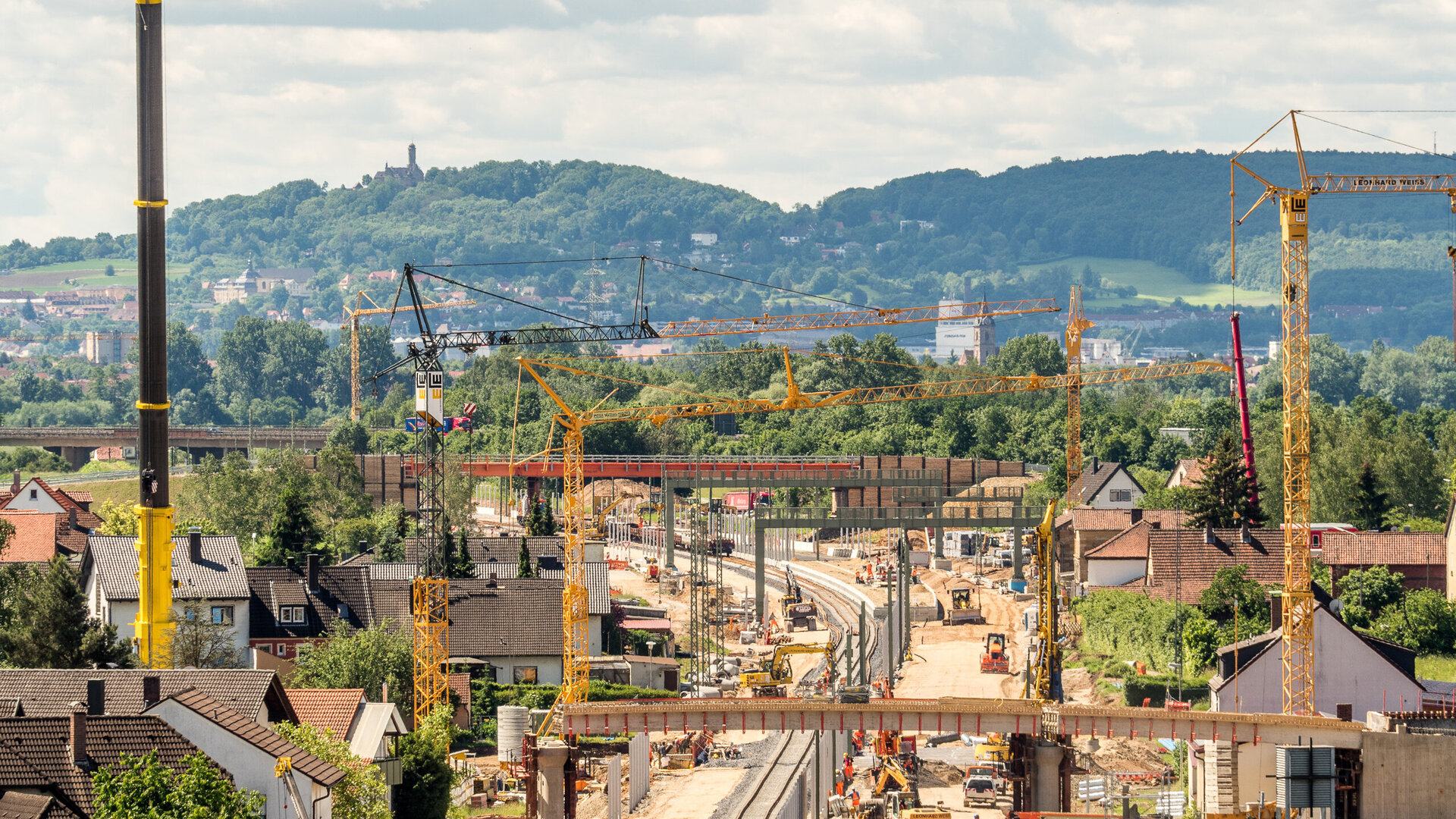 The scene shows a bustling construction site with cranes, vehicles and new tracks, surrounded by residential buildings and nature.