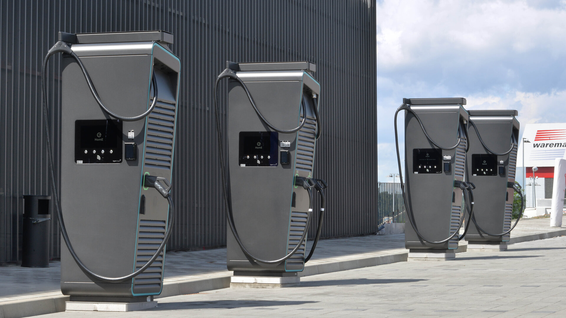 Vier moderne Ladestationen für Elektroautos stehen auf einem gepflasterten Platz, umgeben von einer hohen Wand.