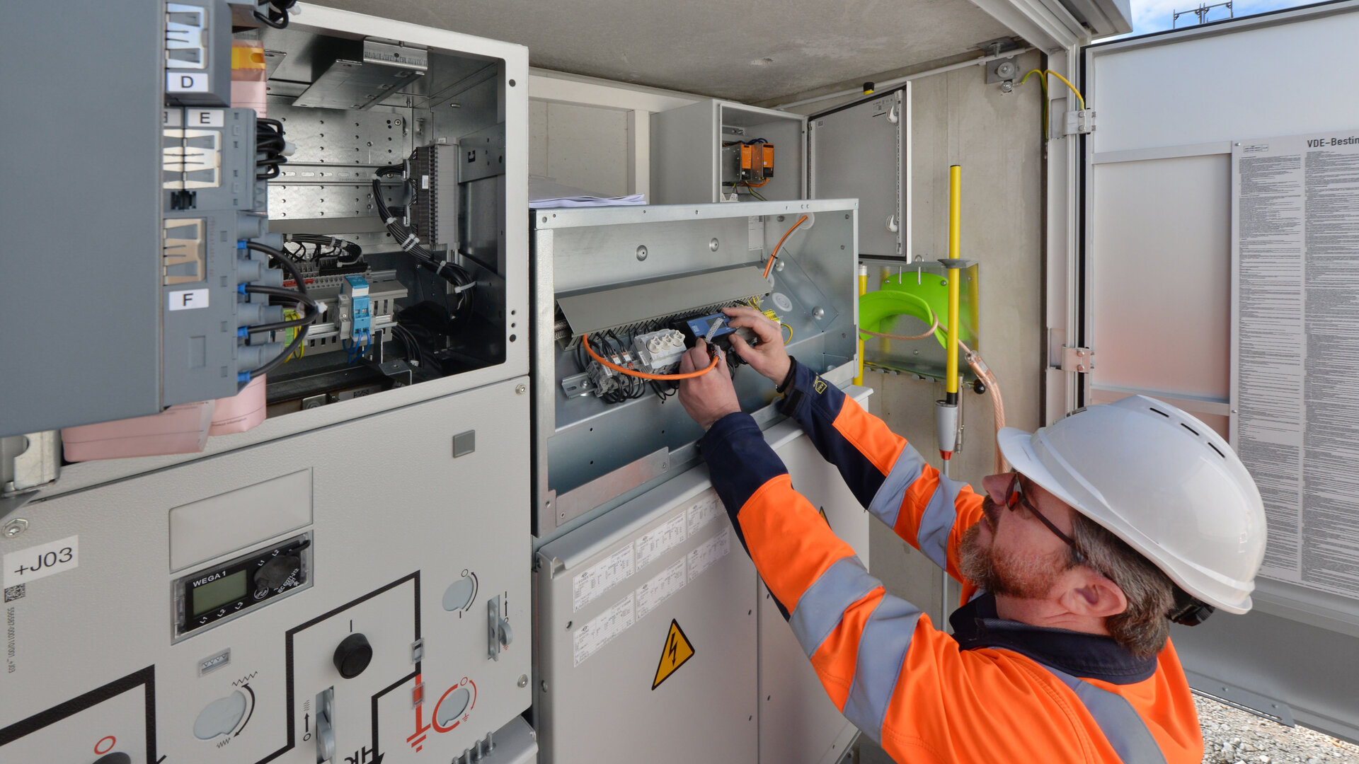Ein Elektriker mit Schutzhelm und Warnjacke arbeitet an einem offenen Schaltschrank im Freien. Er prüft Kabelverbindungen und Bauteile innerhalb der Anlage. Im Hintergrund sind weitere Schaltschränke, Beschriftungen und ein blauer Himmel sichtbar.