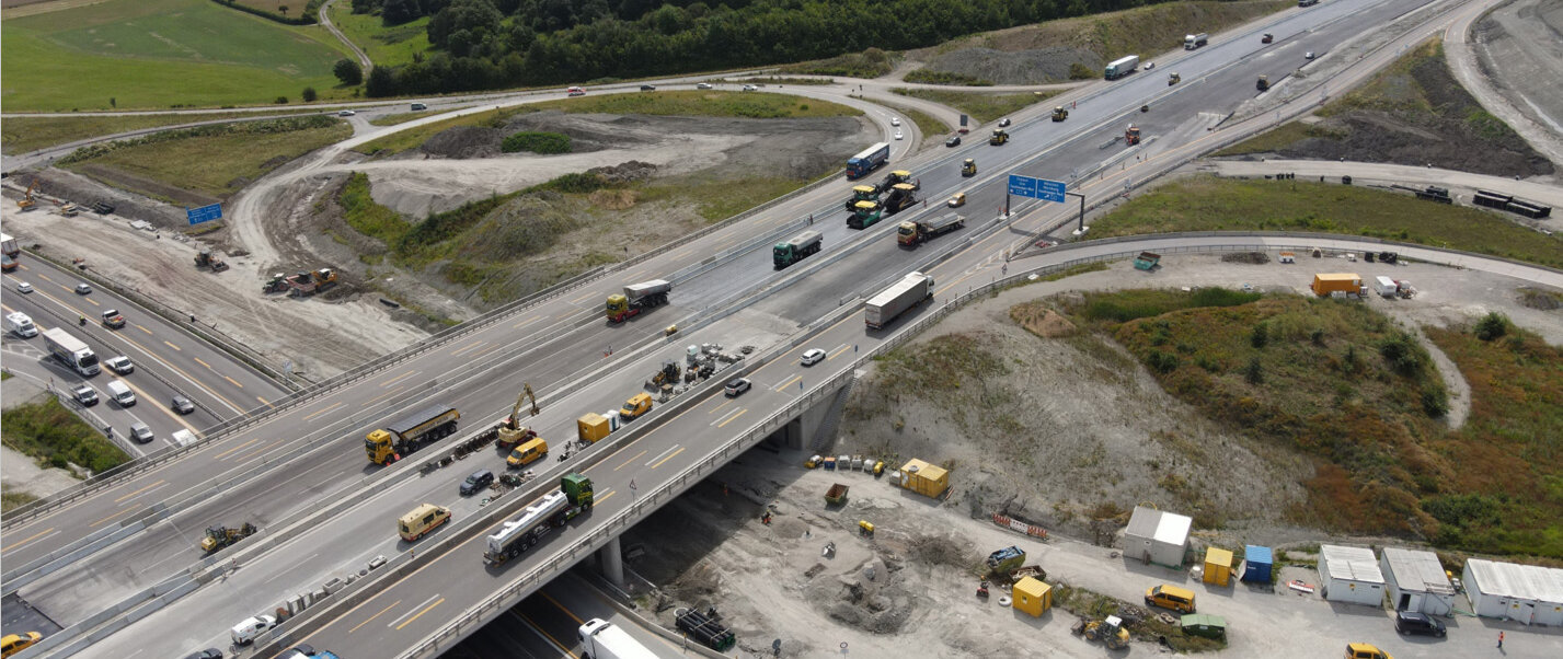 Eine Luftaufnahme zeigt eine vielbefahrene Baustelle mit mehreren Baustellenfahrzeugen und Lkw auf einer Autobahn.