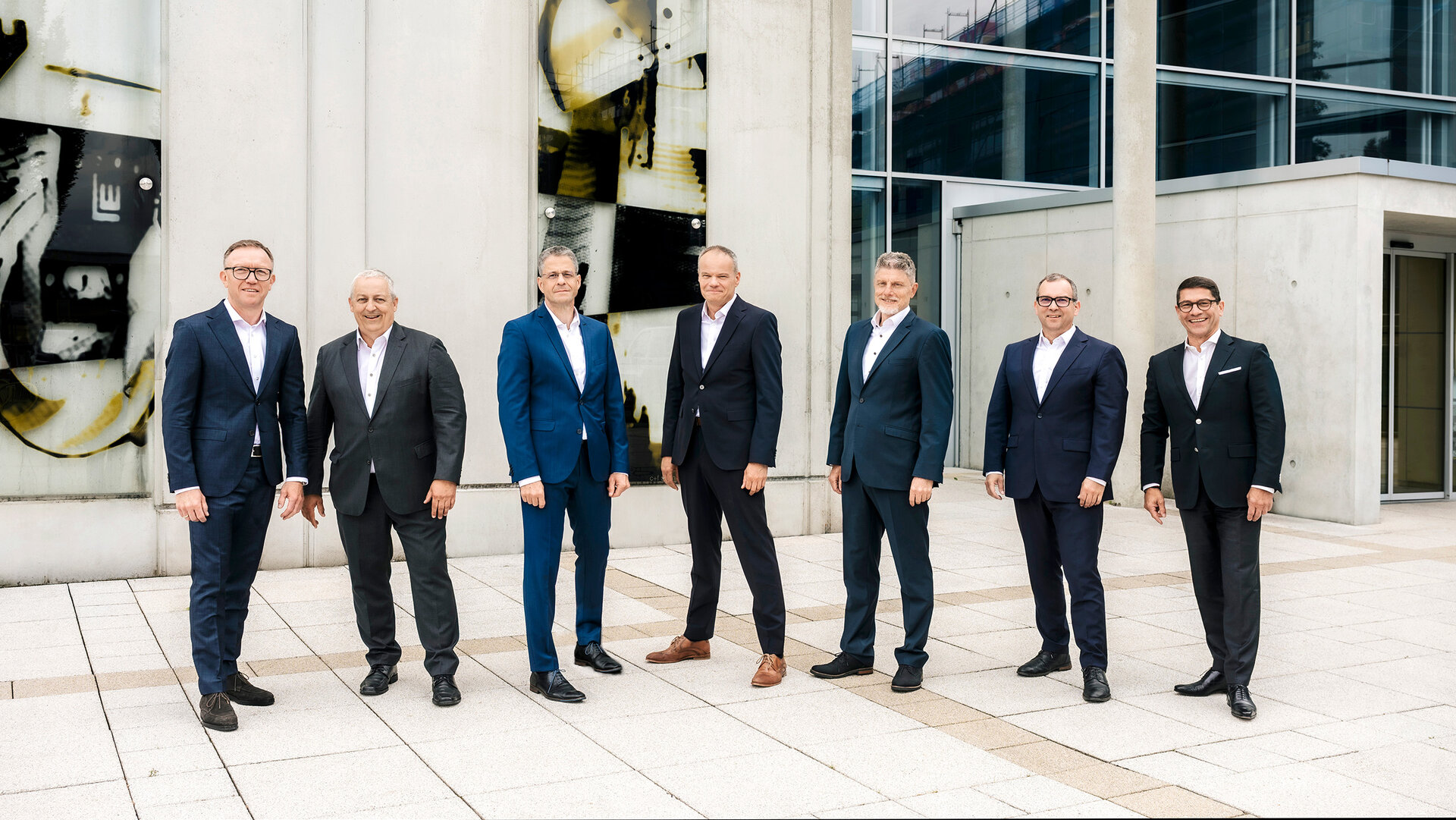Gruppenbild der Geschäftsführung in Anzügen stehen im Freien vor einem modernen Bürogebäude.