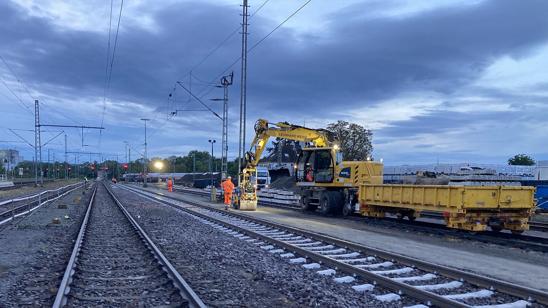 Im Dämmerlicht arbeiten Bauarbeiter und ein Zweiwege-Bagger beim Erstellen des Randwegs entlang der Gleise.