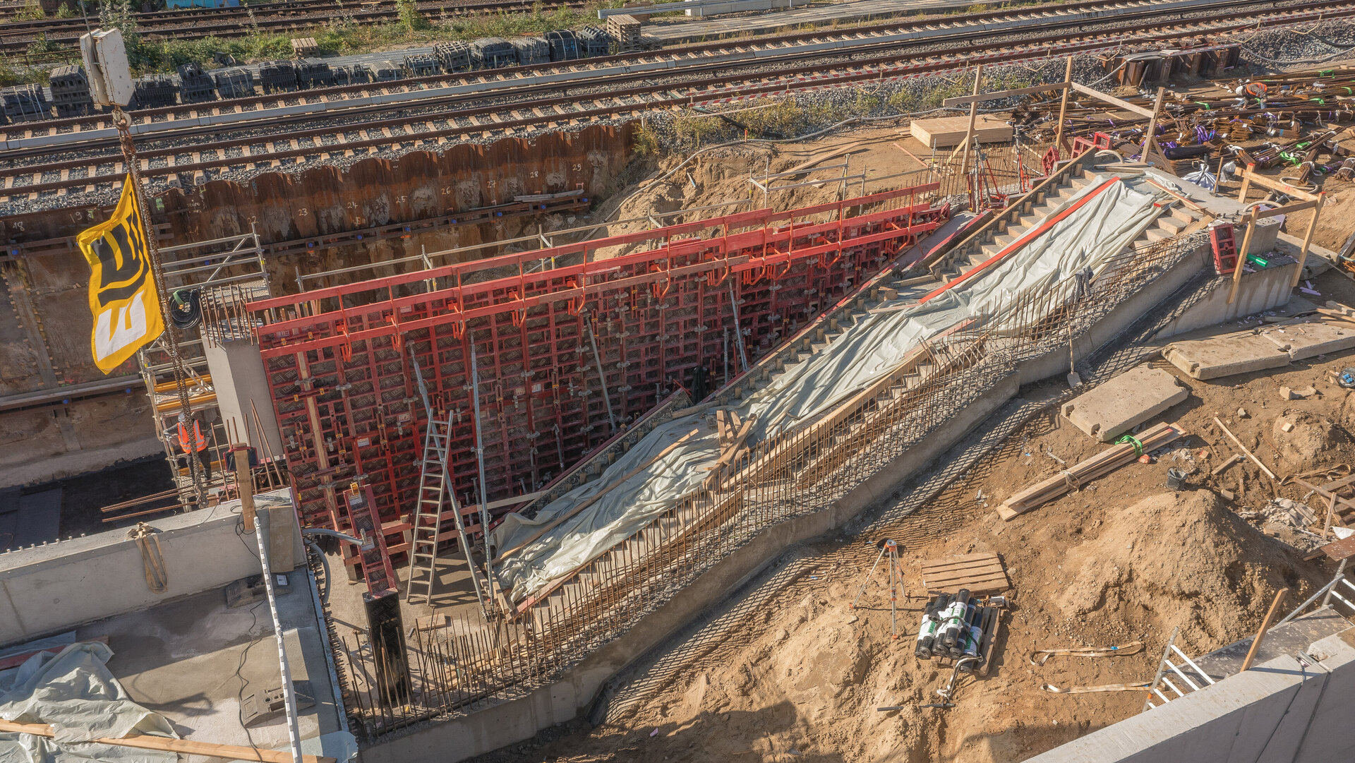 Eine Baustelle mit einem großen, roten Schalungsgerüst, das für den Bau einer Rampe vorbereitet ist, umgeben von Erde und Werkzeugen.