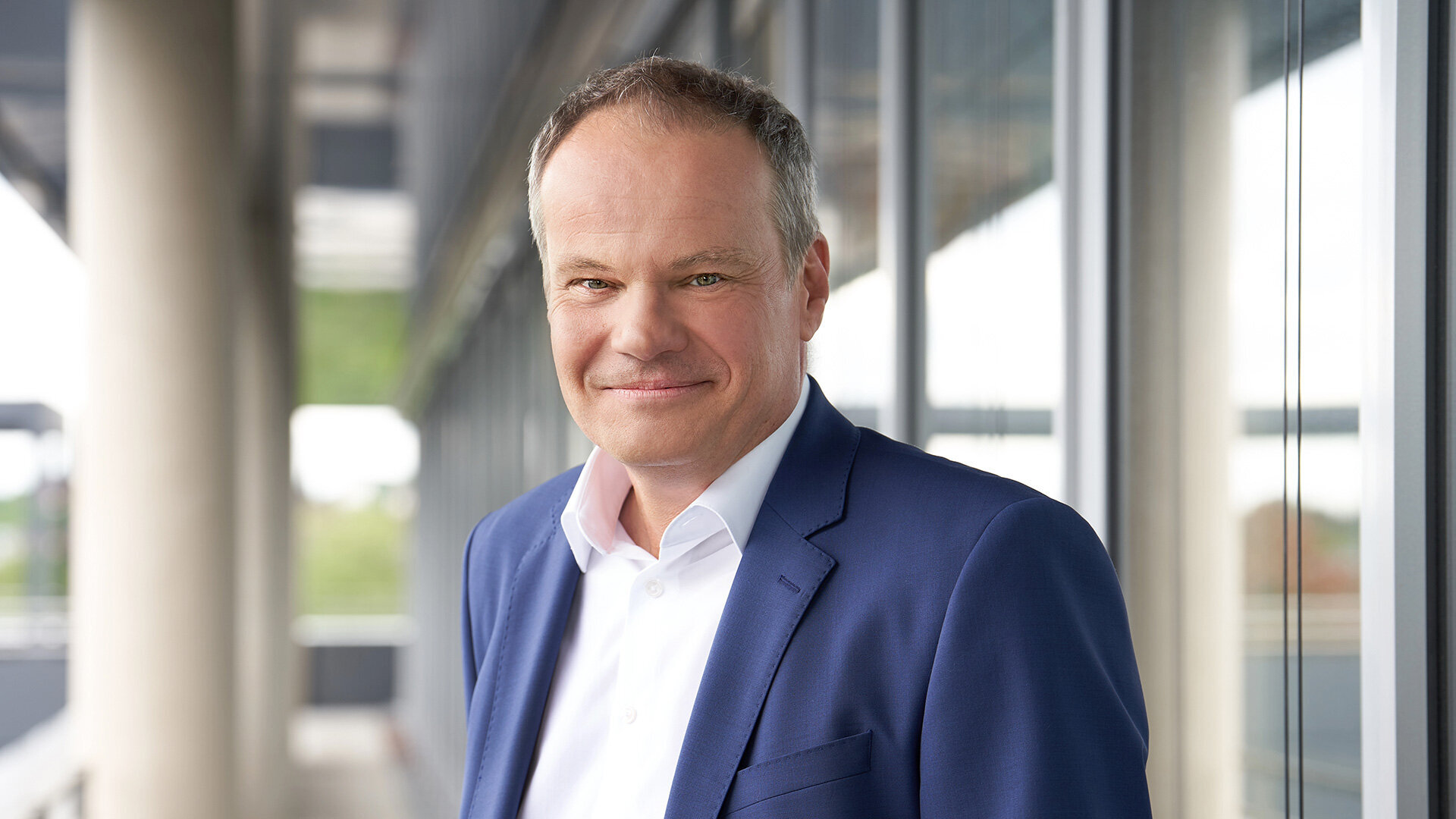 Foto von Christian Ott, Ressortleiter Unternehmenssteuerung und stellvertretender Vorsitzender der Geschäftsführung, stehend in Anzug und Hemd vor einem modernen Bürogebäude.