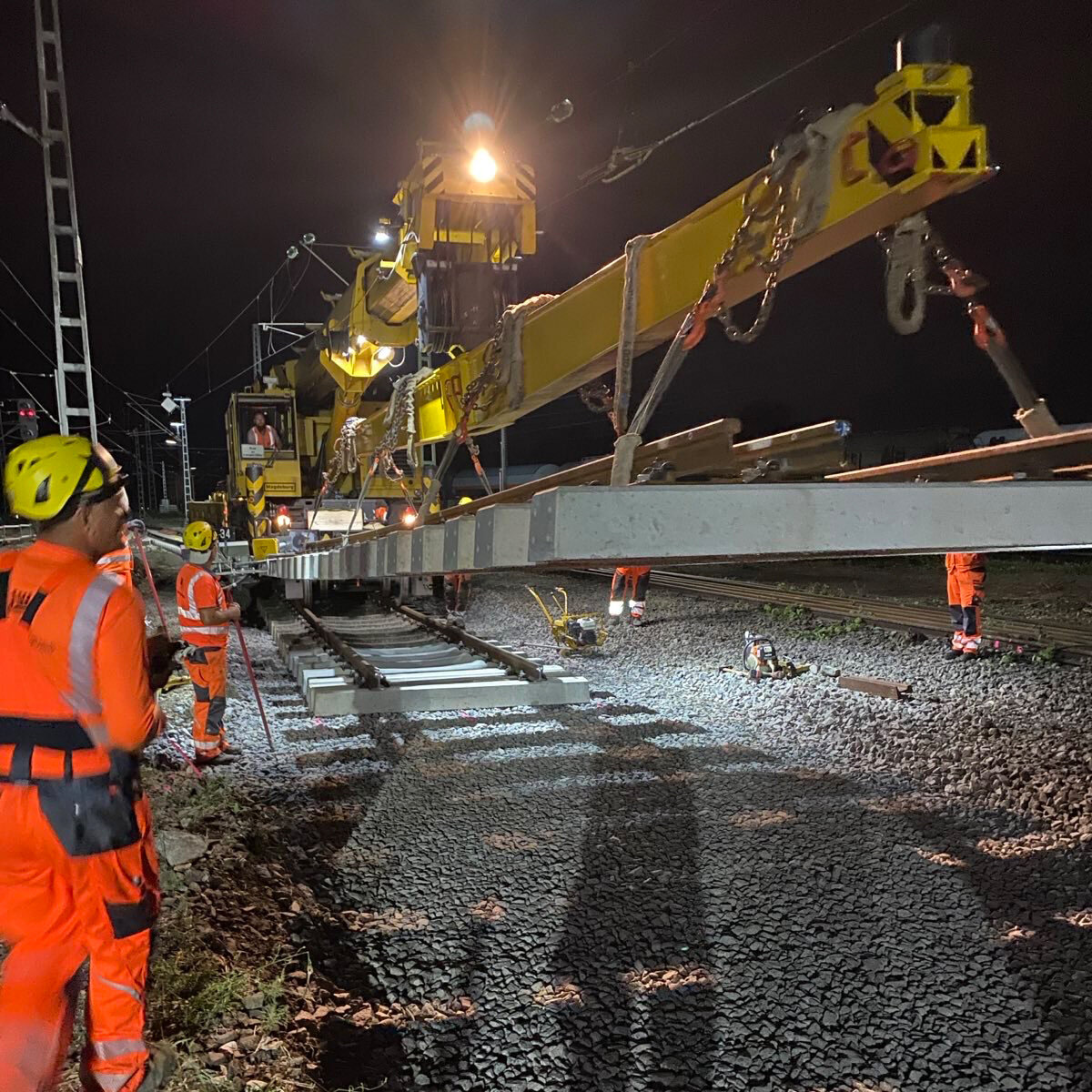 Bei Nacht arbeiten mehrere Bauarbeiter in orangefarbener Kleidung am Weicheneinbau mittels Schienenkran.