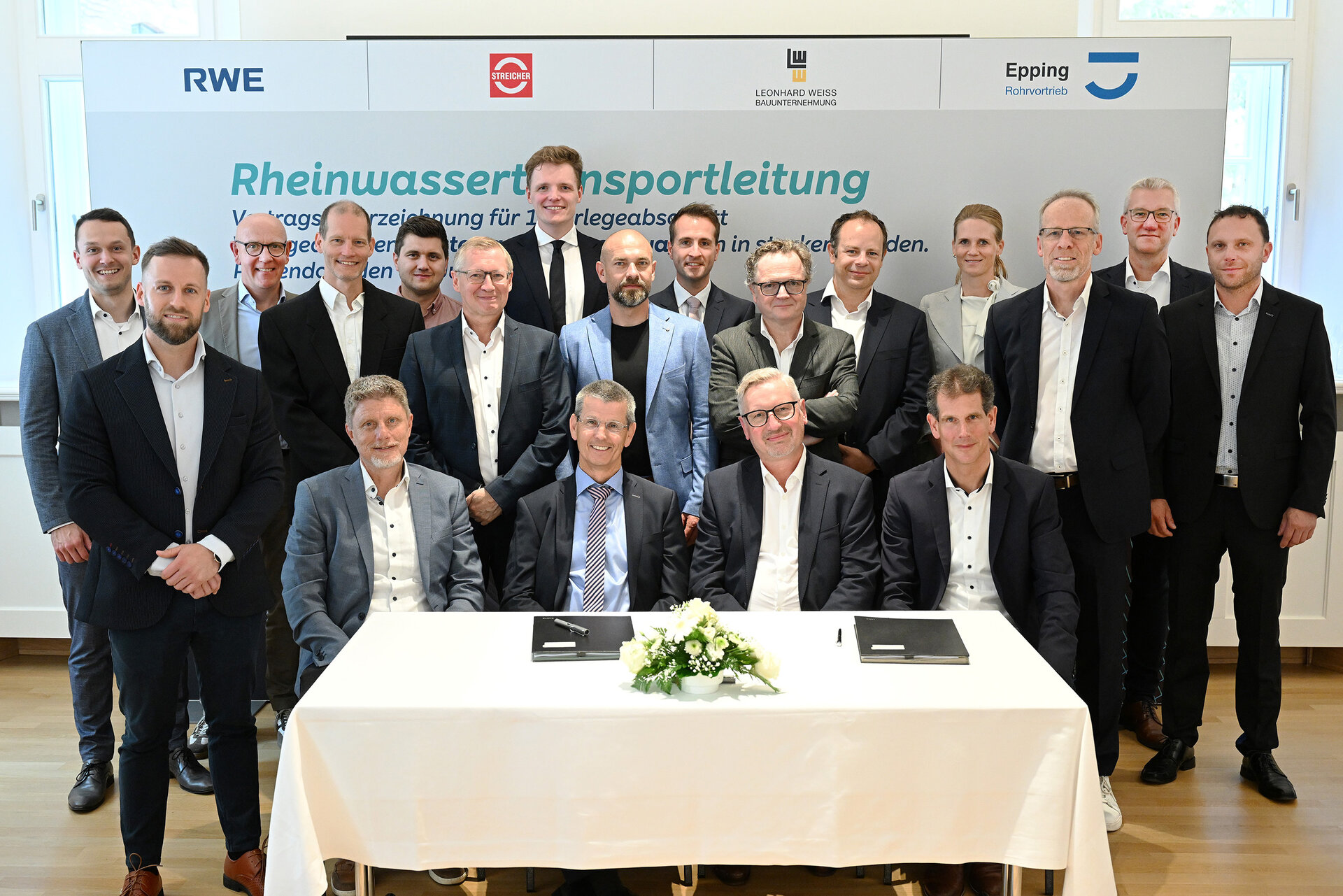 Gruppenfoto bei der Vertragsunterzeichnung zur Rheinwassertransportleitung: Vertreter von RWE, Streicher, Leonhard Weiss und Epping Rohrvortrieb posieren gemeinsam. Vorne sitzen vier Männer am Tisch mit unterschriftsbereiten Dokumenten, im Hintergrund stehen weitere Beteiligte in Anzügen und Businesskleidung.