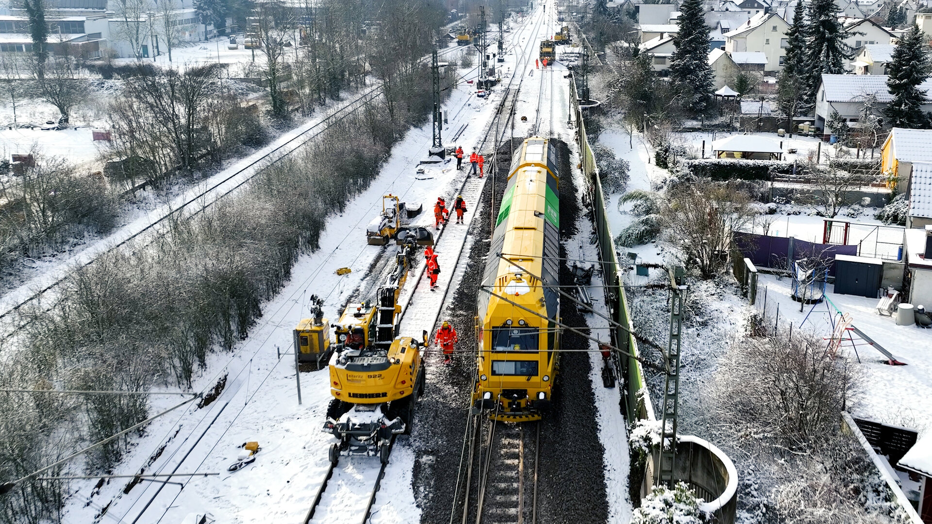 Luftaufnahme eines gelben Gleisbaufahrzeug neben einem Zug und eine Gruppe von Arbeitern,die Gleisarbeiten in einer schneebedeckten Umgebung durchführen.