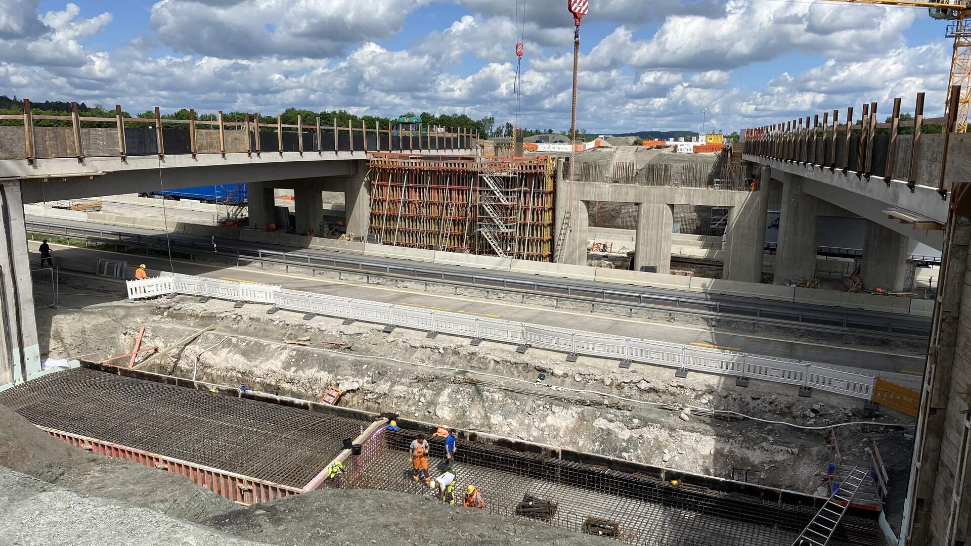 Großbaustelle einer Brücke über eine Autobahn: Bauarbeiter in orangefarbener Schutzkleidung arbeiten an Bewehrungselementen im Fundamentbereich. Im Hintergrund sind Kräne, Schalungen und bereits errichtete Betonpfeiler und Fahrbahnteile sichtbar. Der Himmel ist blau mit einigen Wolken.