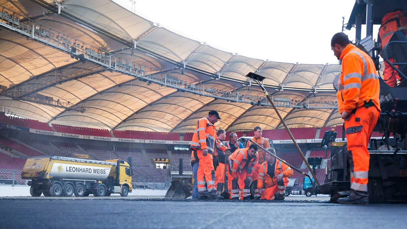 In einem Stadion arbeiten mehrere Bauarbeiter in orangefarbener Kleidung an der Asphaltierung des Spielfelds.
