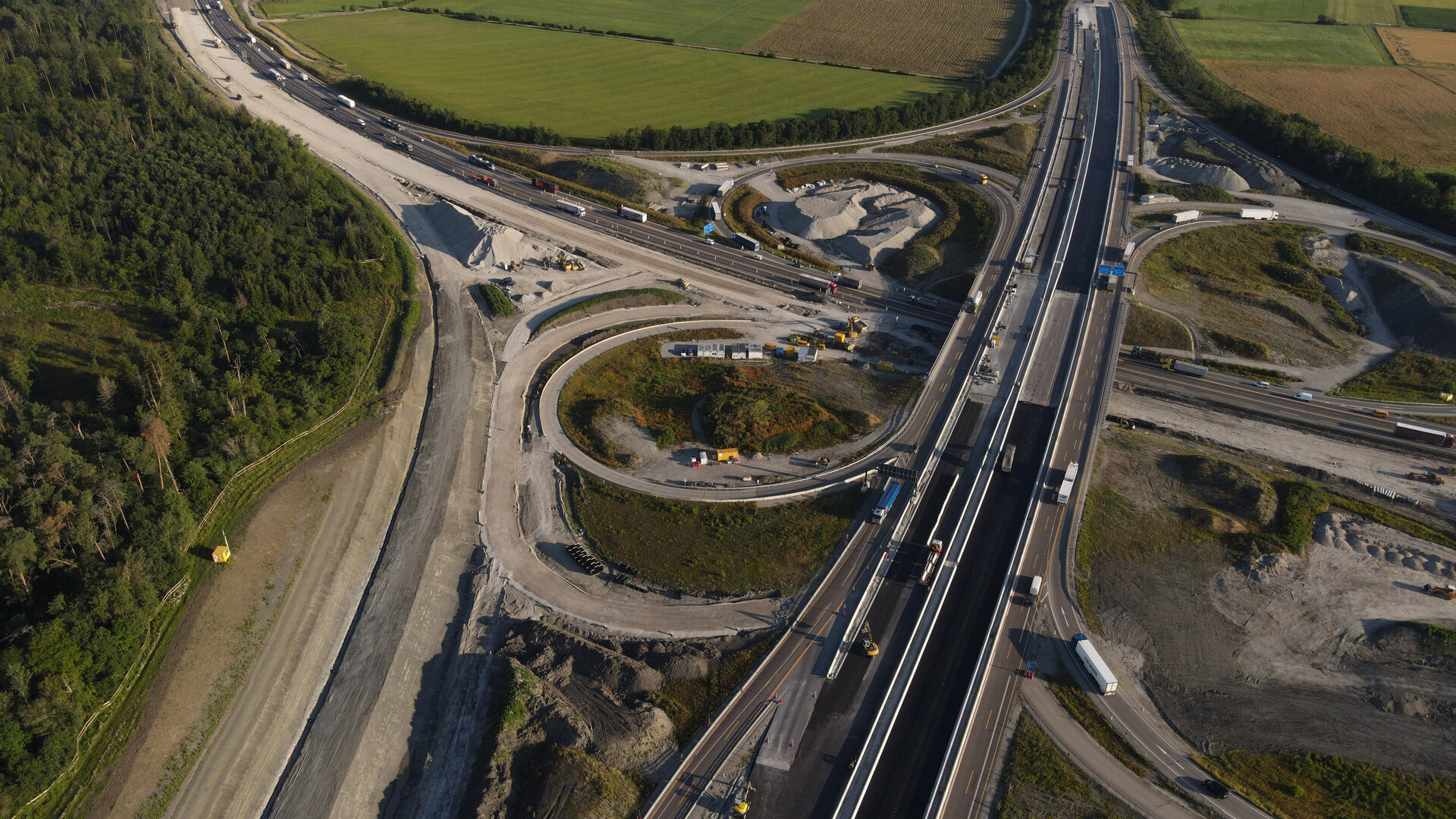 Die Landschaft zeigt eine Baustelle mit verschiedenen Straßenführungen, umgeben von grünen Wiesen und einem Wald im Hintergrund.